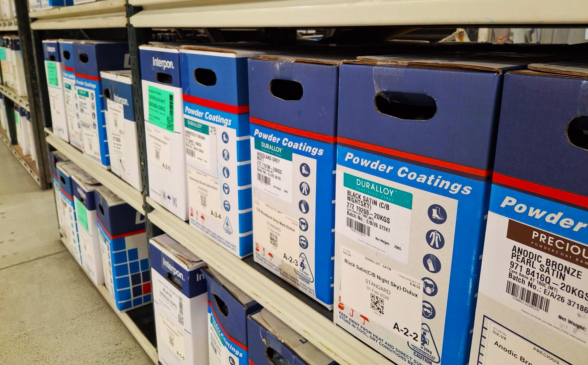 Boxes of powder coating paint stored on warehouse shelving, showing a range of colour finishes used for security screen frames.